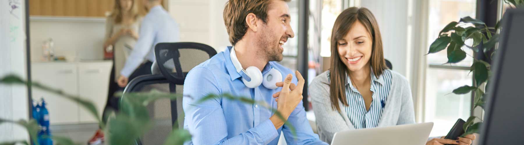 Two colleagues sit at a desk next to a laptop having a discussion.