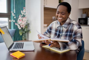 Individual engaged in online learning with laptop and holding book.